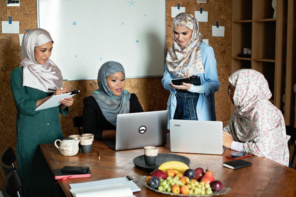 Four Muslim businesswomen in hijabs working together with laptops in a modern office setting.